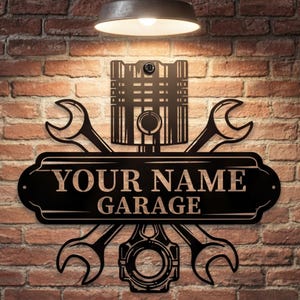 Può includere: Insegna da garage in metallo nero con la scritta "YOUR NAME GARAGE". L'insegna presenta chiavi incrociate, un pistone e altri elementi a tema automobilistico, montata su un muro di mattoni.