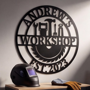 May include: A circular black metal wall sign featuring the text ANDREW'S WORKSHOP and EST. 2023, with silhouettes of a hammer, wrench, saw blade, and other tools integrated into the design. The sign is mounted on a wall above a black tool chest.