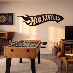 May include: A black Hot Wheels wall sign featuring the brand logo in a stylized flame shape. The sign is mounted on a light-colored wall above a wooden foosball table with black legs, positioned in a room with a dartboard and a television.