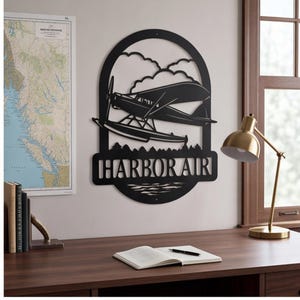 May include: A black metal wall art piece featuring a seaplane flying above water with clouds in the background. The design is enclosed in an arched frame with the text HARBOR AIR cut out in a bold, sans-serif font at the bottom of the structure.