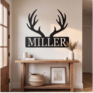 以下が含まれることがあります： 太いセリフ体のフォントで「MILLER」という文字が刻まれた長方形のネームプレートの上に、鹿の角が配置された黒い金属製のウォールサイン。壁に取り付けられたこのサインは、取り付け穴のあるすっきりとしたダークなシルエットデザインが特徴です。
