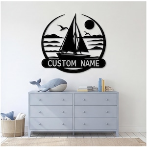 May include: A circular black metal wall sign featuring a sailboat silhouette on water with birds and a sun. The design includes a horizontal banner at the bottom with the text CUSTOM NAME in a sans-serif font, framed by a curved border.