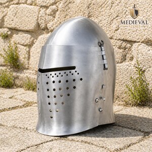 May include: A polished, silver-colored medieval helmet with a rounded top and a visor. The helmet features a series of small holes for ventilation and a hinged section on the side. The Medieval Store logo is visible in the background.