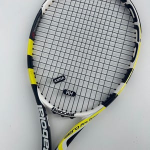 May include: A Babolat Aero Pro Drive tennis racket featuring a white, black, and yellow frame with black strings. The racket includes two small rectangular labels on the strings with the text ANNA and RH printed in white on a black background.