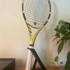 Könnte beinhalten: Ein gelb-schwarzer Babolat-Tennisschläger mit weißem Griff, ausgestellt auf einem schwarzen Ständer. Der Schläger hat ein schwarzes Saitenmuster und das Babolat-Logo ist auf dem Rahmen sichtbar. Der Ständer steht auf einer Holzoberfläche.