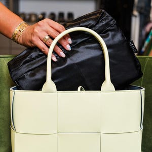 Könnte beinhalten: Eine hellgelbe Handtasche mit gewebtem Design und gebogenem Griff. Eine schwarze Satintasche befindet sich in der Handtasche. Die Tasche steht auf einem grünen Samtstuhl.