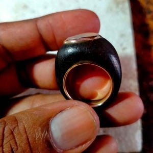 Puede incluir: Anillo artesanal con una banda de madera oscura, interior dorado y una piedra o incrustación de color claro en la parte superior. El anillo se sostiene en una mano, mostrando su superficie lisa y pulida. Diseño moderno.