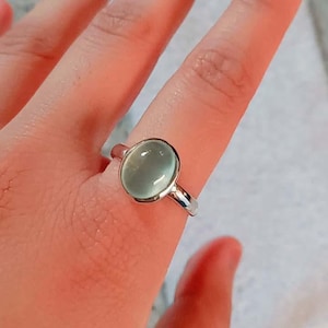 May include: A silver ring with a large, oval, pale green gemstone. The gemstone is set in a polished silver band. The ring is a piece of jewellery.