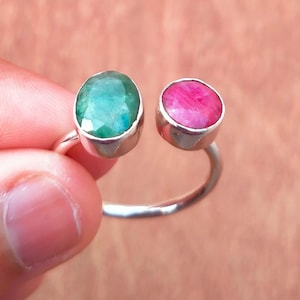 Puede incluir: Un anillo de plata con dos piedras preciosas ovaladas: una verde esmeralda y otra magenta. Las piedras están engastadas en un bisel. El anillo tiene un diseño abierto.