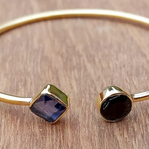 May include: Gold-toned open cuff bracelet with a blue square gemstone and a black round gemstone. The bracelet is set against a wooden surface, showcasing the jewelry's design and color contrast.