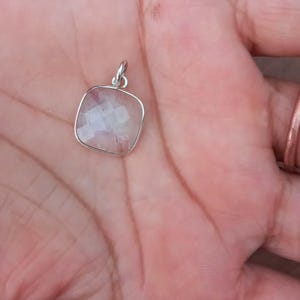 Può includere: Un ciondolo quadrato con una gemma sfaccettata e iridescente incastonata in una montatura in metallo color argento. Il ciondolo è mostrato su uno sfondo color carne.