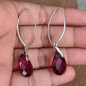 May include: A pair of silver-toned drop earrings featuring faceted, teardrop-shaped, deep red gemstones. The earrings have a simple, elegant design with a curved hook that goes through the ear. The gemstones are a rich, dark red color.
