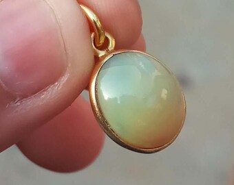 May include: A gold-colored pendant with an oval, iridescent gemstone. The gemstone has a pearly, opalescent appearance with a mix of pale green and yellow hues. The pendant is held up by a small gold loop.