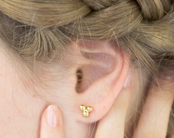 May include: A close-up of a person's ear with a small gold earring featuring three tiny balls. The person is wearing a gold ring on their finger.