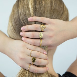 May include: A woman's hand wearing five gold rings. The rings are all different styles, including a textured band, a thin band, a ring with a small flower design, a ring with a small oval design, and a ring with a large oval design.