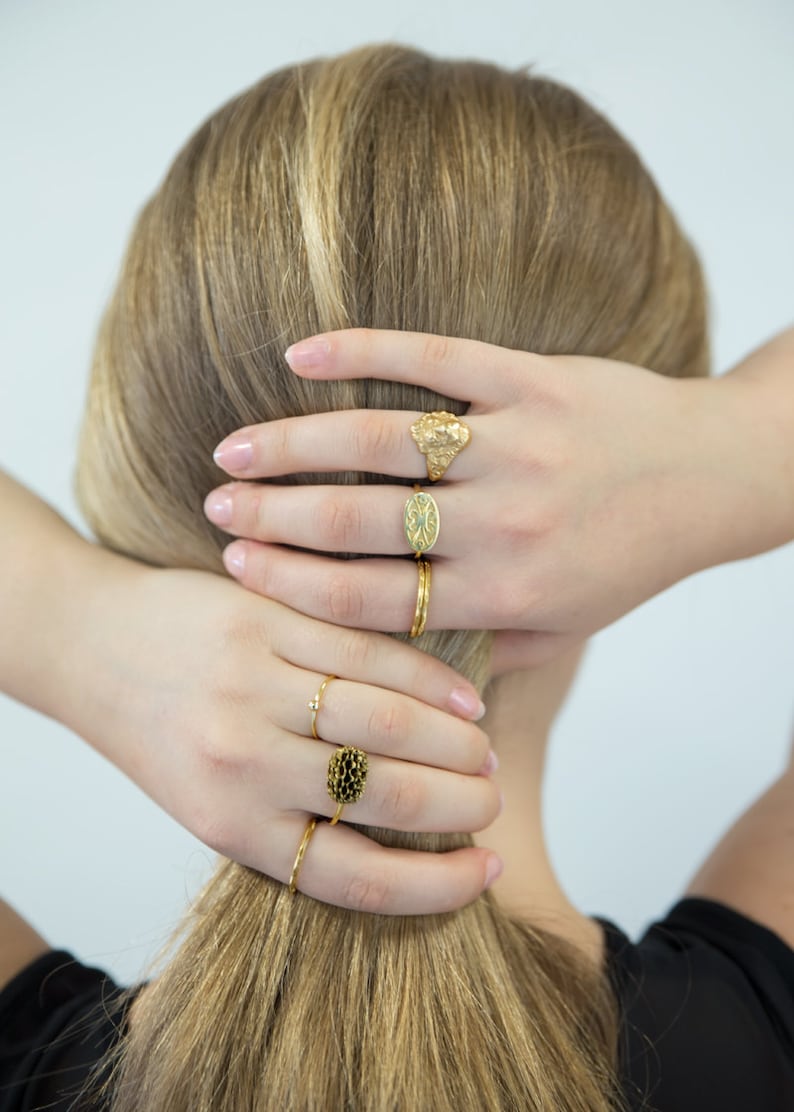 May include: A woman's hand wearing five gold rings. The rings are all different styles, including a textured band, a thin band, a ring with a small flower design, a ring with a small oval design, and a ring with a large oval design.