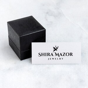 May include: A black square box with a textured surface and a white card with the text "SHIRA MAZAR JEWELRY" and a stylized deer logo.