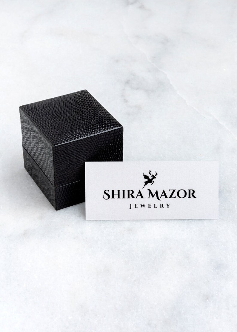 May include: A black square box with a textured surface and a white card with the text "SHIRA MAZAR JEWELRY" and a stylized deer logo.