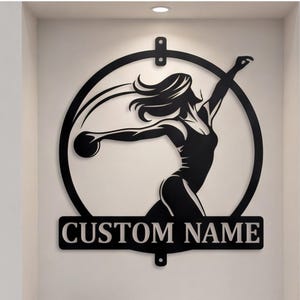 Op de afbeelding: Zwarte metalen wandkunst met een silhouet van een vrouwelijke discuswerper in een cirkelvormig frame. De tekst "CUSTOM NAME" staat eronder. Een paar grijze en blauwe sportschoenen staan op een plank eronder.