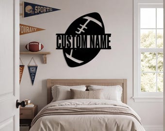 May include: A black metal wall sign shaped like an American football with a horizontal bar across the centre featuring the text CUSTOM NAME in a bold, sans-serif font. The sign is mounted on a light-coloured wall above a bed with neutral bedding.