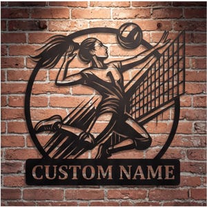 Puede incluir: Arte de pared de metal negro que representa a una jugadora de voleibol en pleno remate, con un balón y una red. La jugadora está dentro de un marco circular. Las palabras "CUSTOM NAME" se muestran en una placa rectangular debajo. El fondo es una pared de ladrillos.