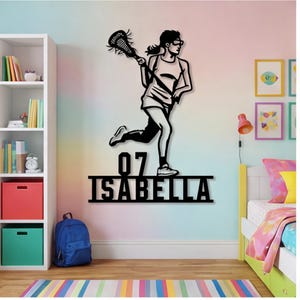Może przedstawiać: Czarna metalowa grafika ścienna przedstawiająca zawodniczkę lacrosse w akcji, trzymającą kij do lacrosse. Dzieło sztuki zawiera numer "07" i imię "ISABELLA" pod postacią. Grafika ścienna jest zamontowana na ścianie z kolorowym gradientem.