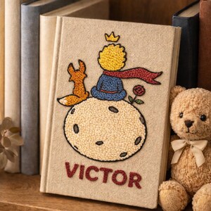 May include: A beige book featuring an embroidered design of a crowned character, a fox, and a rose. The title "VICTOR" is embroidered in red. A teddy bear and other books are visible in the background.