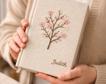 May include: A beige linen journal with a floral embroidery design. The embroidered design features a brown branch with pink and white flowers. The name "Judith" is embroidered below the floral design. The journal has a beige ribbon bookmark.