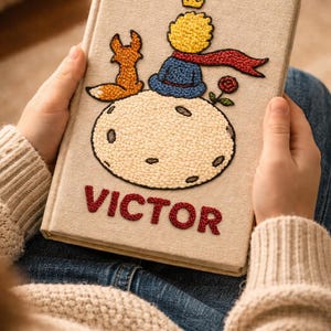 May include: A beige book cover featuring embroidered images of a fox, a prince, and a rose. The word "VICTOR" is embroidered in red. The book is held in a person's hands.