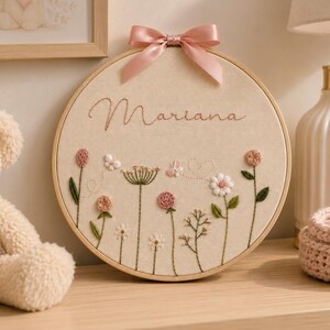 May include: A circular wooden embroidery hoop containing beige fabric with the name Mariana stitched in pink cursive thread. The design features various embroidered wildflowers in shades of pink, white, and green, accented by a pink satin ribbon bow.