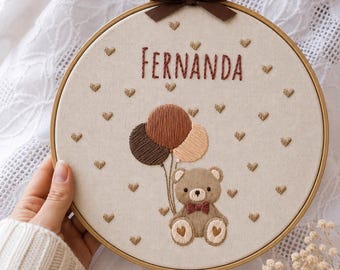 May include: A circular wooden embroidery hoop containing a beige fabric panel with the name FERNANDA stitched in dark red. The design features a brown teddy bear with a red bow tie, three balloons in shades of brown, and small embroidered hearts.