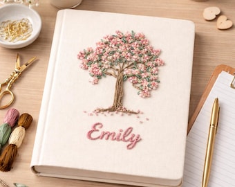 May include: Cream-colored journal with an embroidered tree design in pink and green. The name "Emily" is embroidered below the tree. A gold pen and other stationery items are in the background.