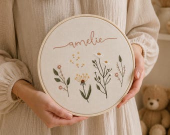 May include: A circular wooden embroidery hoop containing off-white fabric with the name amelie stitched in cursive brown thread above a collection of embroidered wildflowers in shades of yellow, white, green, and pink with delicate stems.