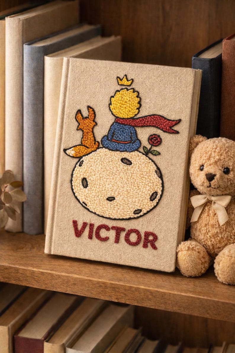 May include: A beige book featuring an embroidered design of a crowned character, a fox, and a rose. The title "VICTOR" is embroidered in red. A teddy bear and other books are visible in the background.