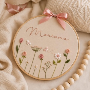 May include: A circular wooden embroidery hoop containing beige fabric with the name Mariana stitched in brown cursive script. The design features embroidered wildflowers in shades of pink, white, and green, accented by a pink satin ribbon bow at the top.