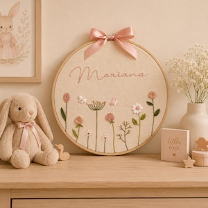 May include: A circular wooden embroidery hoop featuring a cream-colored fabric background with the name Mariana stitched in pink thread. The design includes embroidered pink and white flowers, green stems, and small butterflies, topped with a pink satin bow.