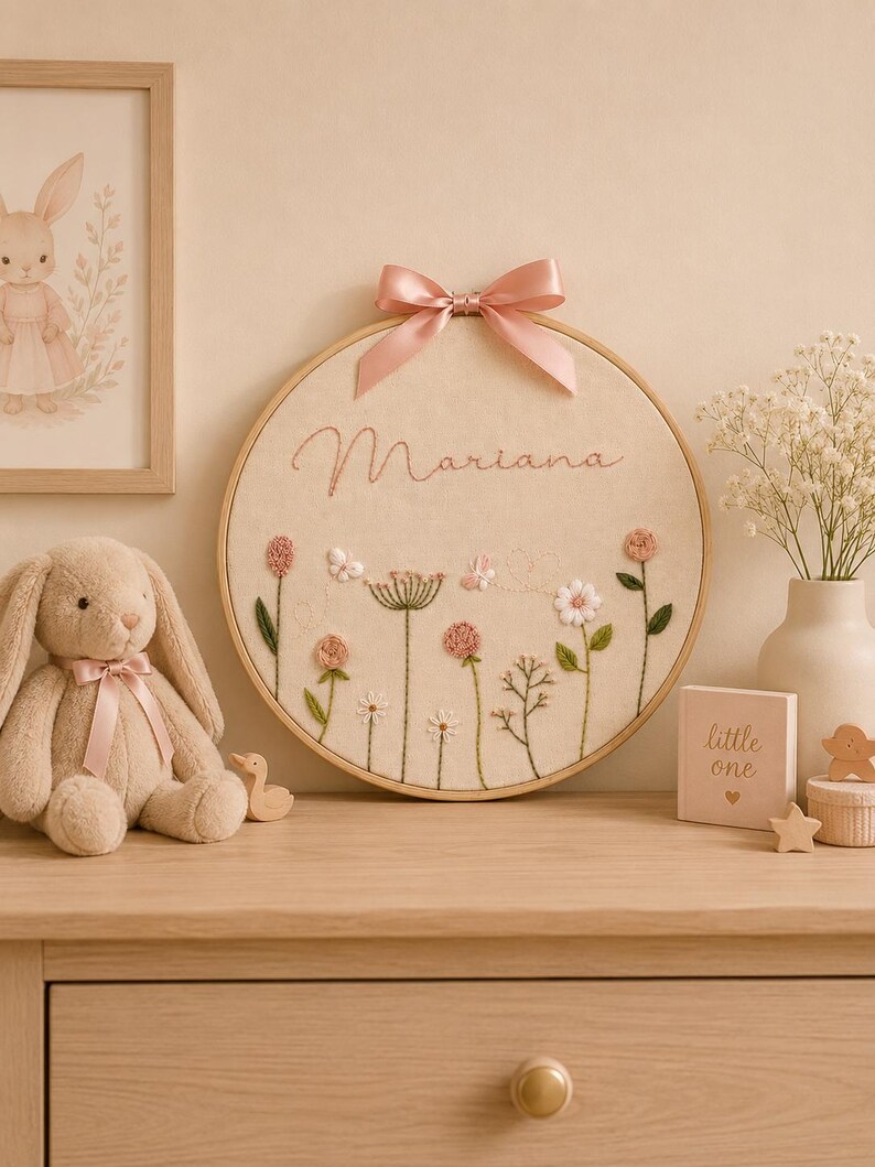 May include: A circular wooden embroidery hoop featuring a cream-colored fabric background with the name Mariana stitched in pink thread. The design includes embroidered pink and white flowers, green stems, and small butterflies, topped with a pink satin bow.
