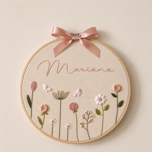 May include: A circular wooden embroidery hoop containing beige fabric with the name Mariana stitched in cursive. The design features embroidered pink and white flowers, green stems, and two small butterflies with a light pink satin ribbon bow.