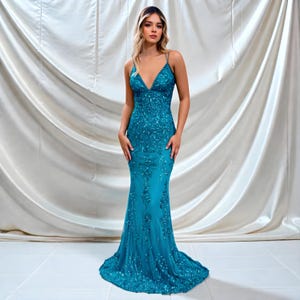 Op de afbeelding: Een turquoise, vloerlange avondjurk met een V-hals en spaghettibandjes. De jurk is versierd met ingewikkeld kralenwerk en pailletten, waardoor een glinsterend effect ontstaat. De rok wijd uit vanaf de knieën en vormt een zeemeerminsilhouet.