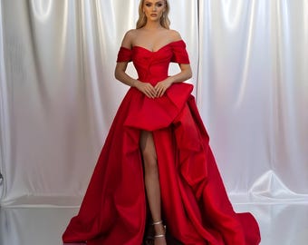 May include: A vibrant red off-the-shoulder gown with a high slit and a dramatic bow detail. The dress features a flowing skirt and is paired with silver heels. The gown's design is elegant and suitable for formal events.