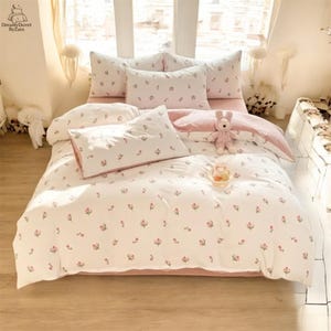 Puede incluir: Un juego de cama blanco con estampado floral y detalles en rosa. El juego incluye una funda nórdica, fundas de almohada y una sábana bajera. La funda nórdica y las fundas de almohada presentan un delicado estampado floral rosa. Un conejito de peluche y un plato pequeño con golosinas están sobre la cama.