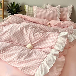 Może przedstawiać: Różowy i biały zestaw pościeli w kratę. Zestaw zawiera poszwę na kołdrę, poduszki i prześcieradło z gumką. Poszwa na kołdrę i poszewki na poduszki mają białe koronkowe wykończenie. Łóżko pokryte jest miękkim, jasnoróżowym prześcieradłem.