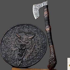 May include: A Viking-style axe with a silver blade and a wooden handle wrapped in leather, next to a round shield with a carved wolf design. The axe and shield are dark brown and black.