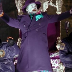 Puede incluir: Una persona con un abrigo morado de doble botonadura, sombrero a juego y pantalones a cuadros, con maquillaje blanco y lápiz labial rojo. La persona lleva un pañuelo verde y turquesa y una flor rosa en la solapa. El fondo incluye decoraciones doradas.