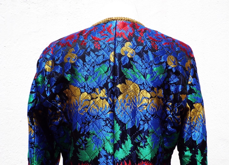 Pode incluir: Um close-up de um casaco com um padr&atilde;o floral vibrante. O casaco apresenta uma base preta com desenhos florais azuis, vermelhos, verdes e dourados. A gola &eacute; adornada com dourado. Pe&ccedil;a vintage ou &uacute;nica.