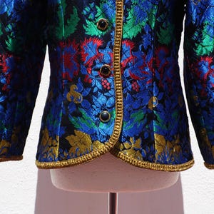 Pode incluir: Casaco colorido com padr&atilde;o floral em azul, verde, vermelho e dourado. O casaco apresenta acabamentos dourados e bot&otilde;es pretos. As mangas s&atilde;o compridas e o tecido tem um aspeto texturizado. Uma pe&ccedil;a de roupa marcante.