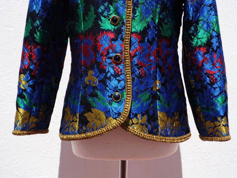 Pode incluir: Casaco colorido com padr&atilde;o floral em azul, verde, vermelho e dourado. O casaco apresenta acabamentos dourados e bot&otilde;es pretos. As mangas s&atilde;o compridas e o tecido tem um aspeto texturizado. Uma pe&ccedil;a de roupa marcante.