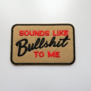 Può includere: Toppa rettangolare beige con testo ricamato in nero e rosso: "SOUNDS LIKE Bullshit TO ME". La toppa ha un bordo nero.