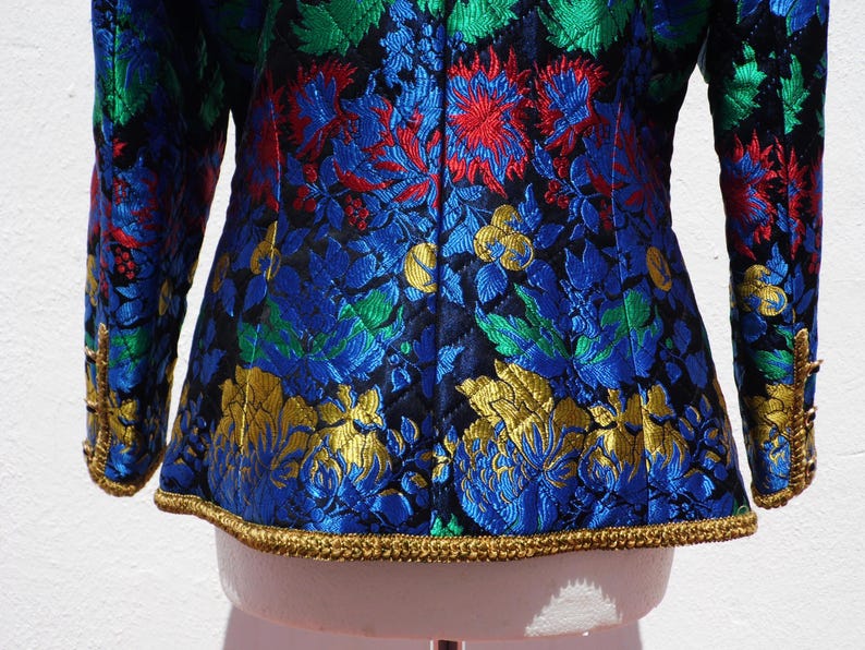 Pode incluir: Um close-up de um casaco colorido com um padr&atilde;o floral. O casaco apresenta uma mistura vibrante de desenhos florais azuis, verdes, vermelhos e dourados sobre um fundo preto. As mangas e a bainha s&atilde;o adornadas com detalhes dourados.
