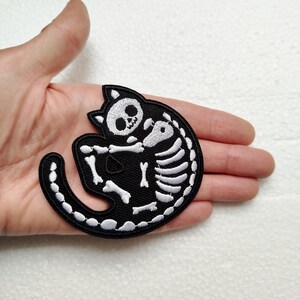 Könnte beinhalten: Schwarz-weißer, gestickter Aufnäher mit einem Katzenskelett-Design. Der Aufnäher zeigt eine zusammengerollte Katze mit ihrem Skelett in Weiß auf schwarzem Hintergrund. Kopf und Schwanz der Katze sind deutlich erkennbar.